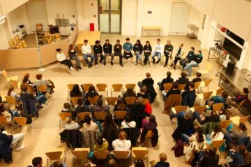 Vogelperspektive, in einer Aula sitzen 12 Personen auf Stühlen dem Publikum, das in mehreren Stuhlreihen sitzt, gegenüber.