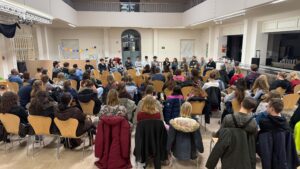 etwa 60 Menschen sitzen in Stuhlreihen und hören einer Gruppe von 14 Personen zu, die ihnen auf Stühlen gegenüber sitzen.