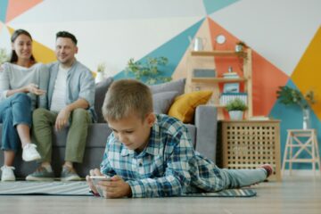 eine Frau und ein mann sitze auf einem Sofa und sehen einem jungen zu, der auf dem Boden liegt und in sein Smartphone guckt.